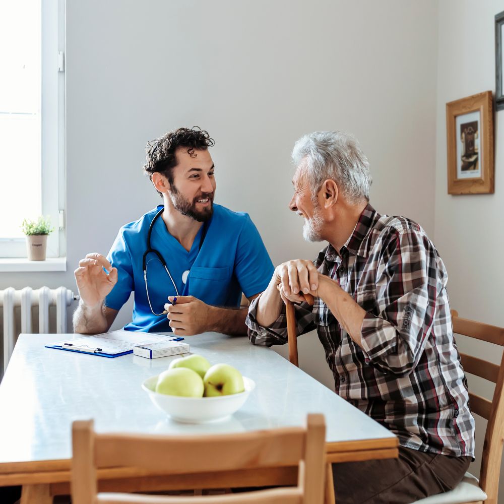 An adult man sits at a table with an elder man with a cane. They appear to be discussing medical care.