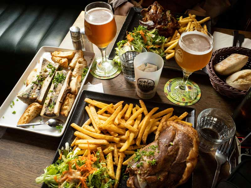 Plates of pub-style food on a table, including sandwiches, fries, roasted meat, salad, bread rolls, and two glasses of beer