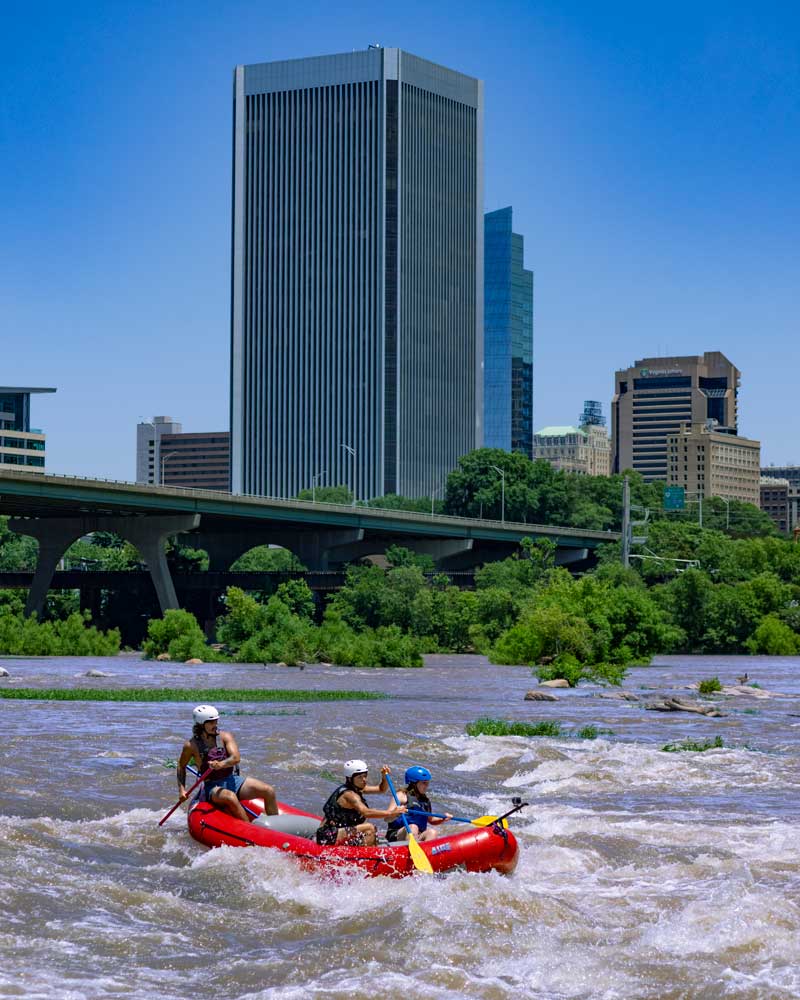 Three people wearing helmets paddle a red raft through rapids on a river with a city skyline and bridge in the background